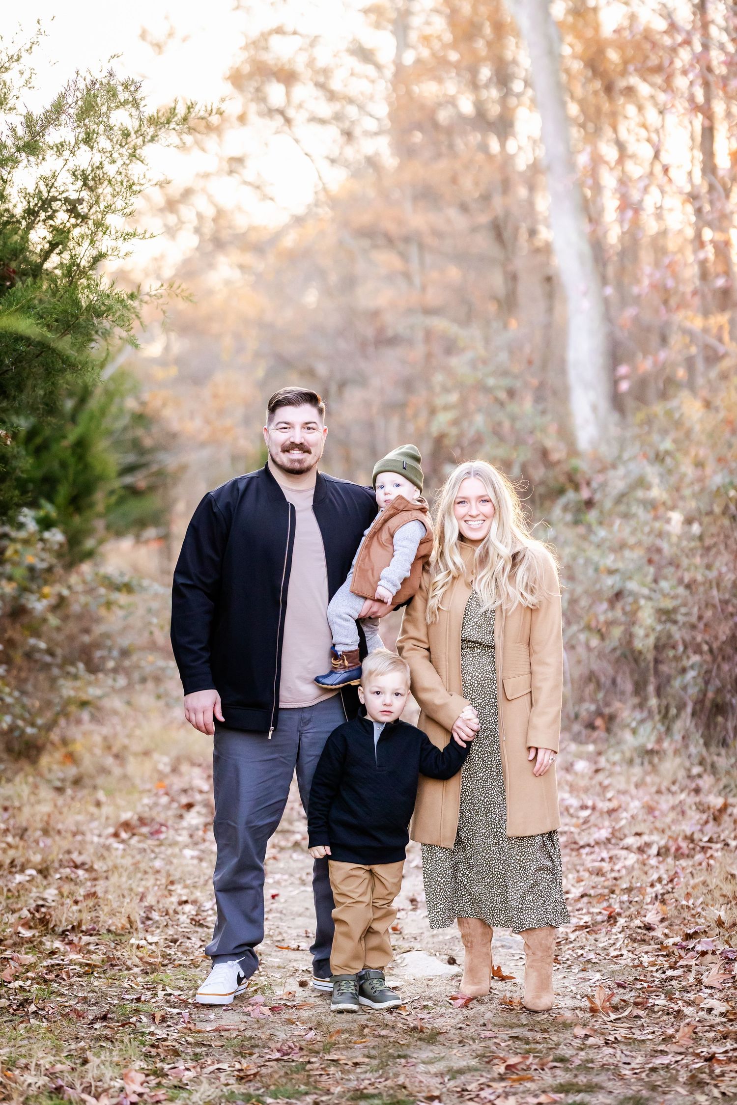 A family in casual fall attire poses together on a leaf-covered forest path during autumn.