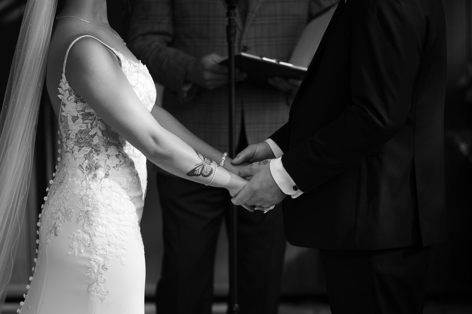 Black and white photo of bride and groom holding hands during wedding ceremony with officiant in background.