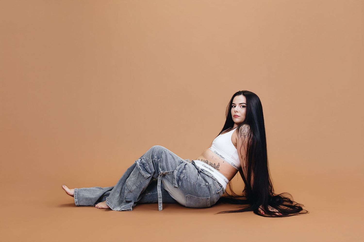 Woman with long black hair sits on floor in white crop top and blue jeans against a warm beige background.