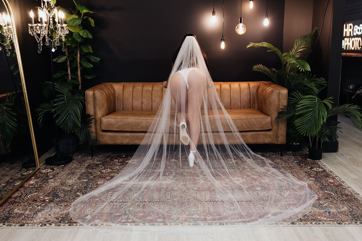 Bride wearing a long white veil in a moody studio setting with leather sofa and tropical plants.