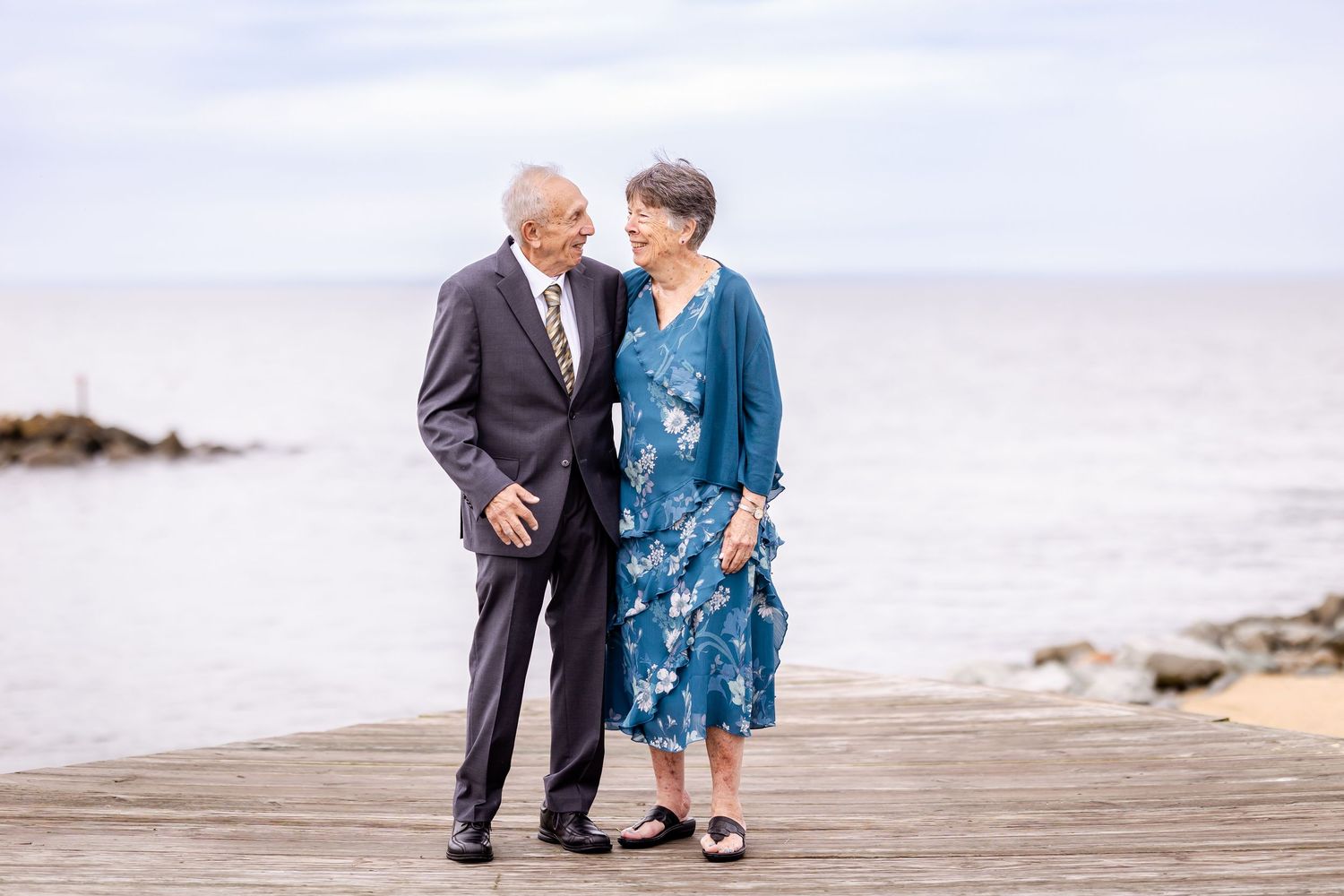 An elderly couple shares a tender moment while walking along a scenic beach boardwalk.