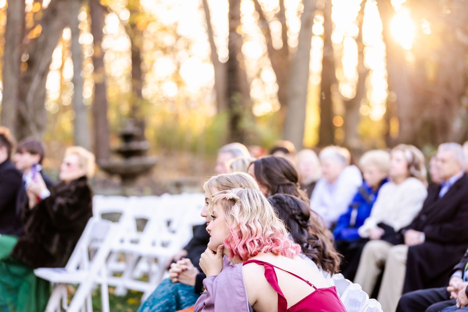 Outdoor wedding ceremony setup in wooded area with white chairs and natural sunlight streaming through trees.