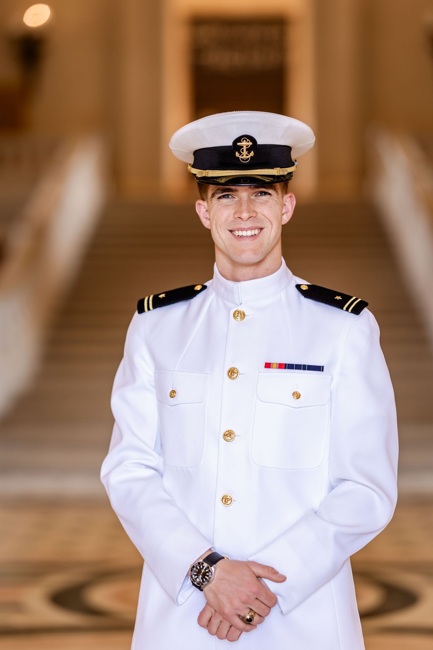 Smiling naval officer in white dress uniform with gold buttons and cap poses indoors.