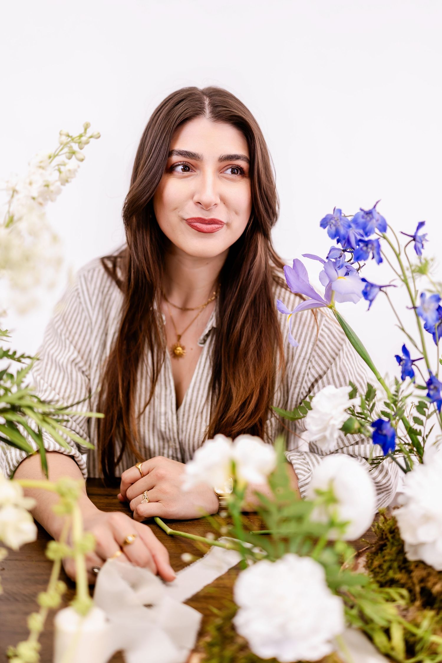 Young woman with long dark hair sits at a wooden table surrounded by white and blue flowers.