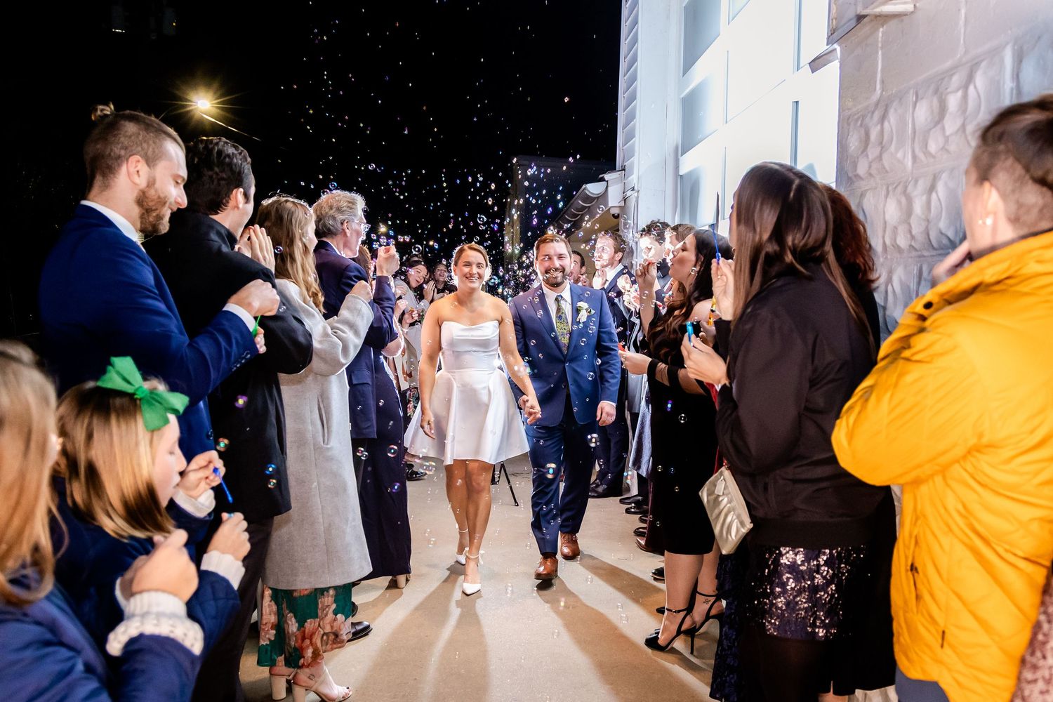 Bride and groom walk through confetti shower surrounded by cheering guests at elegant evening wedding reception exit.
