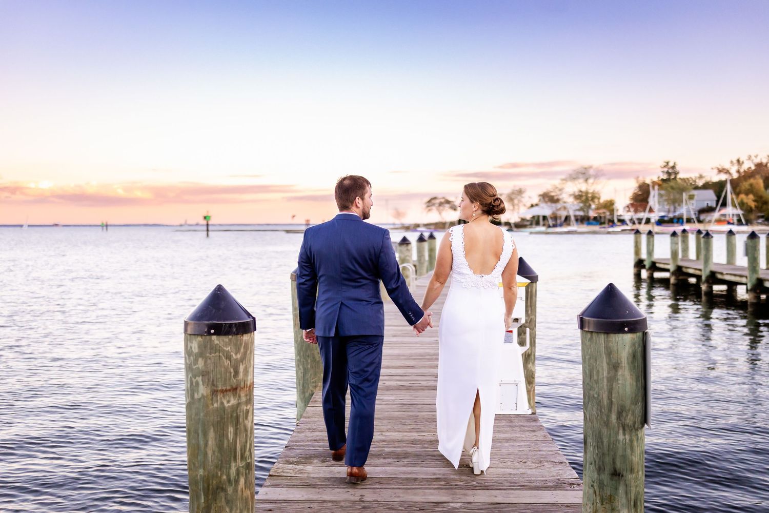 Bride and groom holding hands walking on a waterfront dock at sunset in a scenic coastal wedding setting.