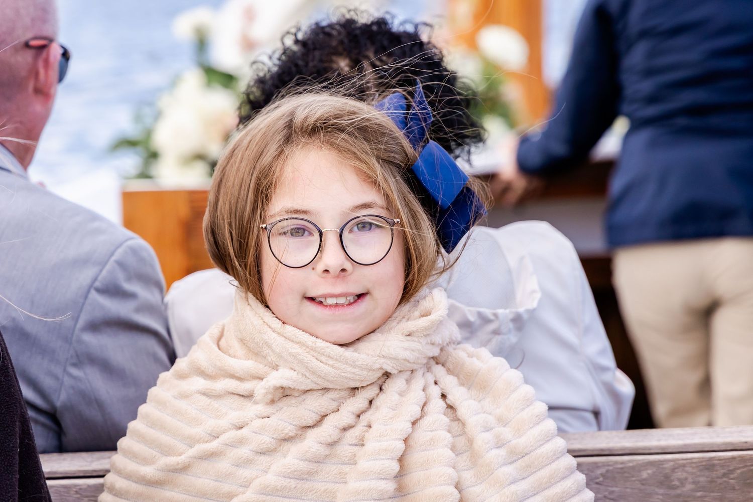 Young girl with glasses and blue hair bow smiles in church pew, wrapped in cream fur shawl at a wedding ceremony.