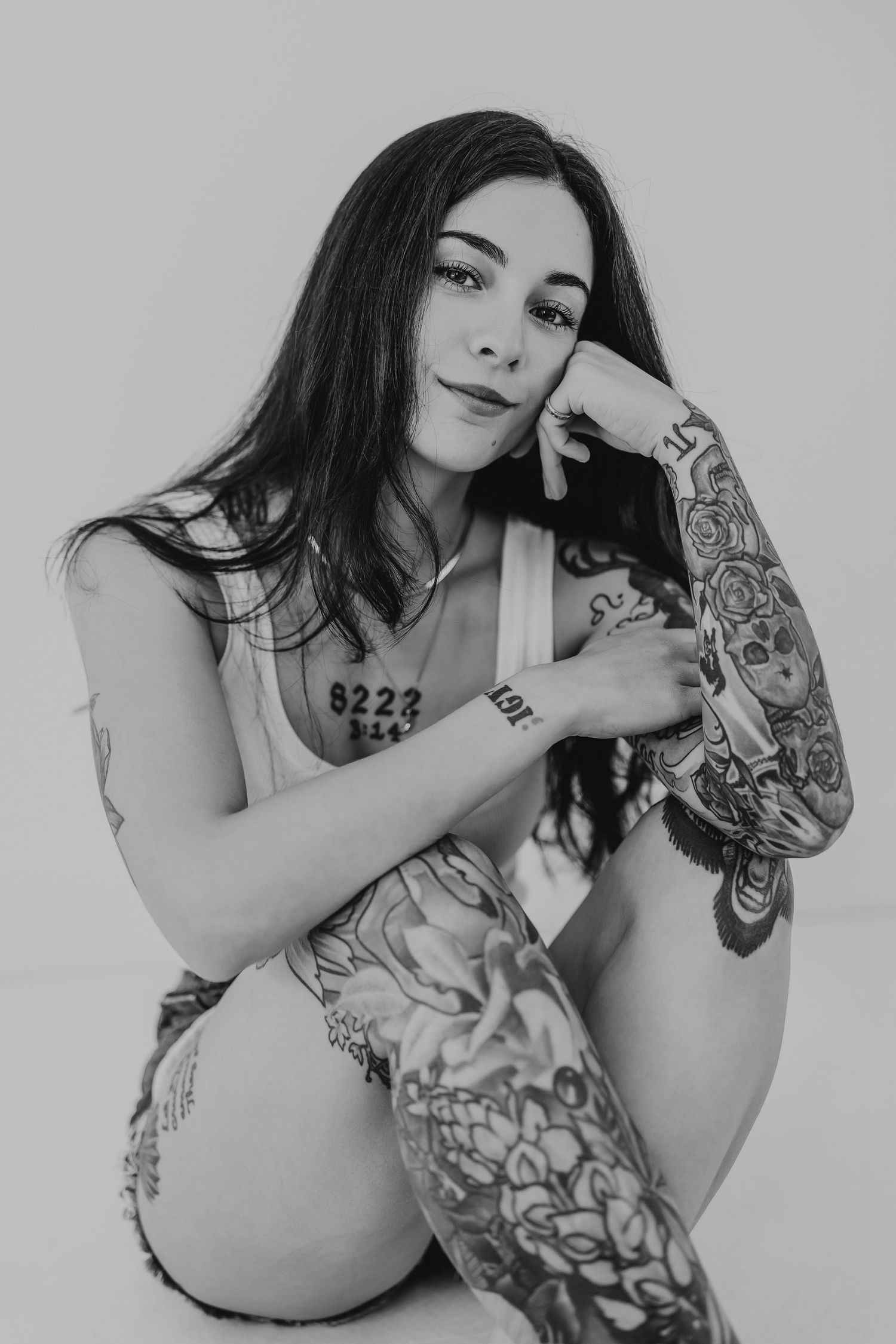 Black and white portrait of a tattooed woman with long dark hair posing confidently against a light background.