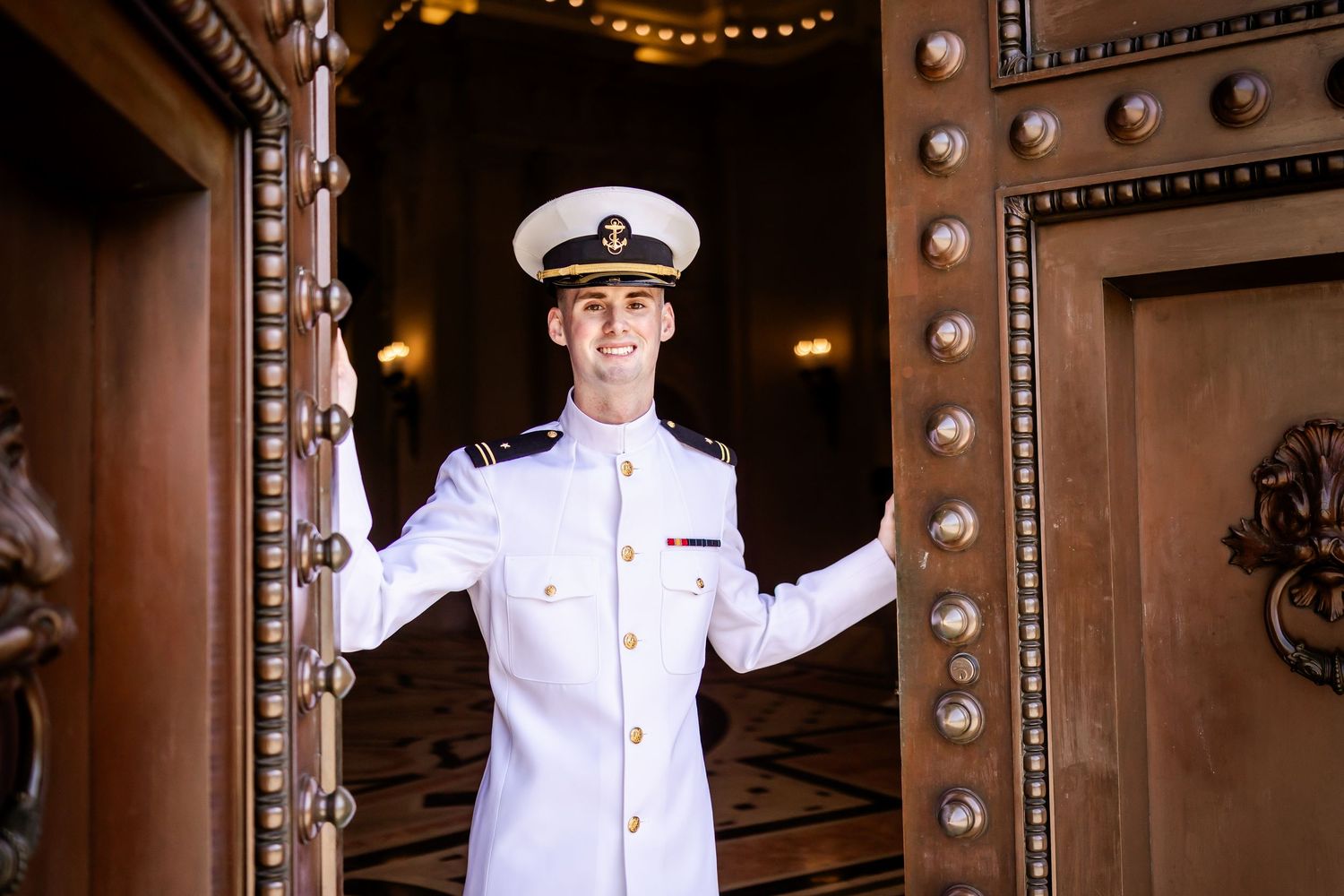 Young naval officer in white dress uniform smiling while holding open a grand ornate wooden door.