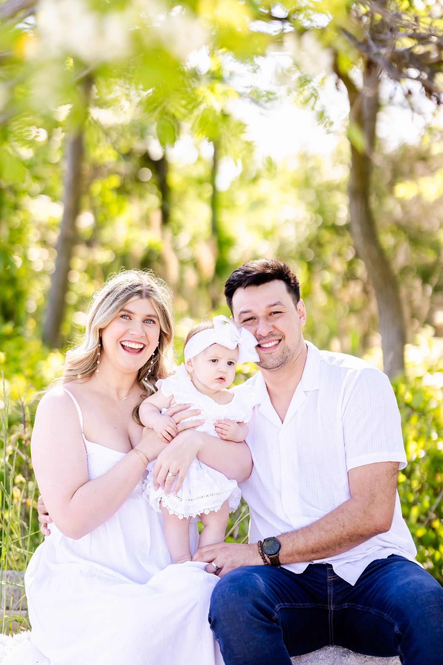 Family in white clothing sits together outdoors on grass surrounded by sunlit trees in a natural setting.