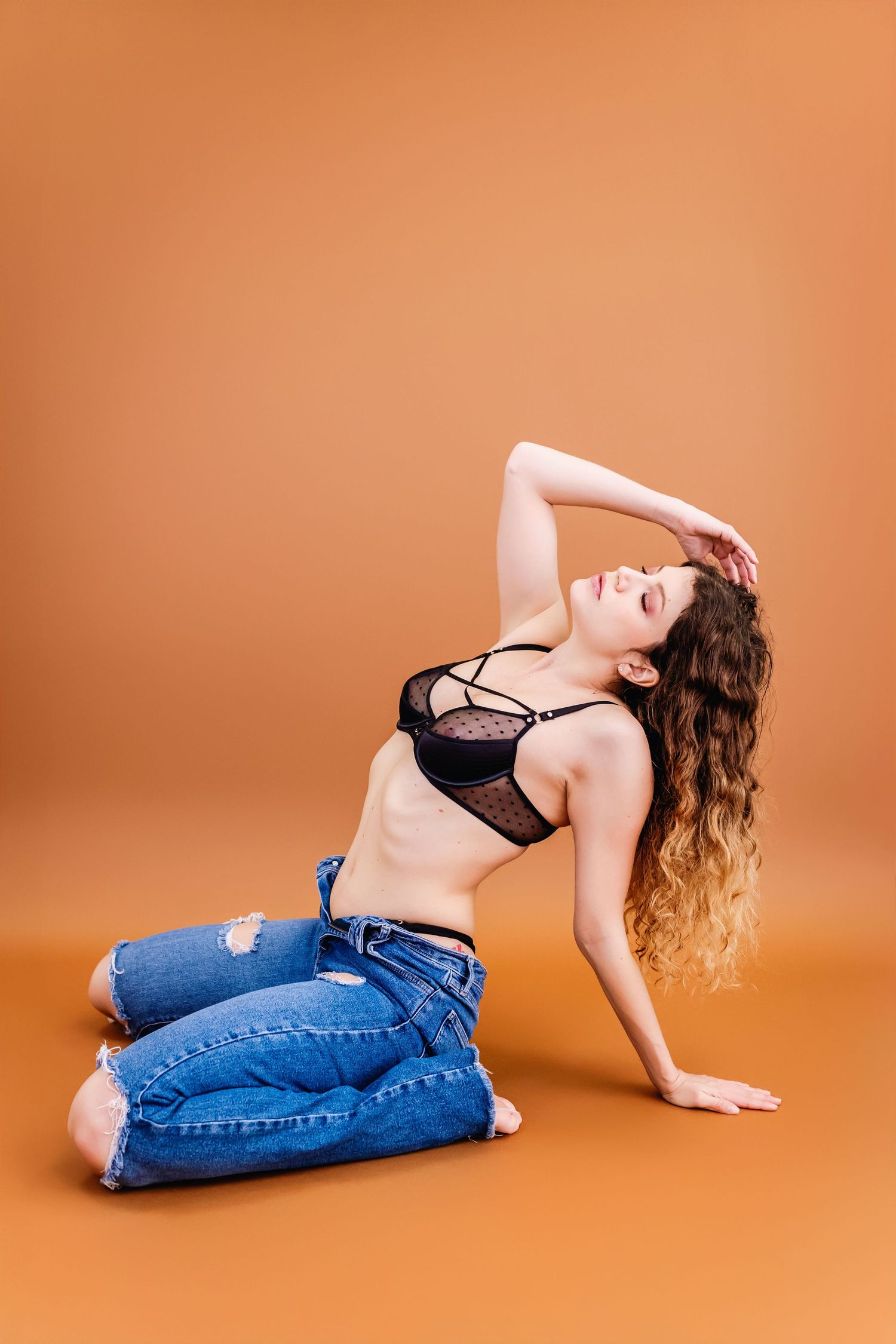 A model in ripped jeans and black lingerie poses artistically against an orange studio backdrop.