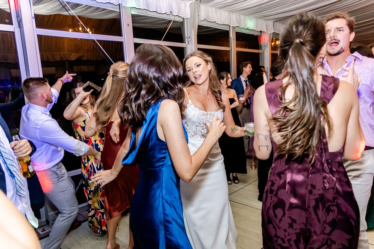 Wedding party dancing and celebrating in a beautifully decorated reception tent with purple lighting.