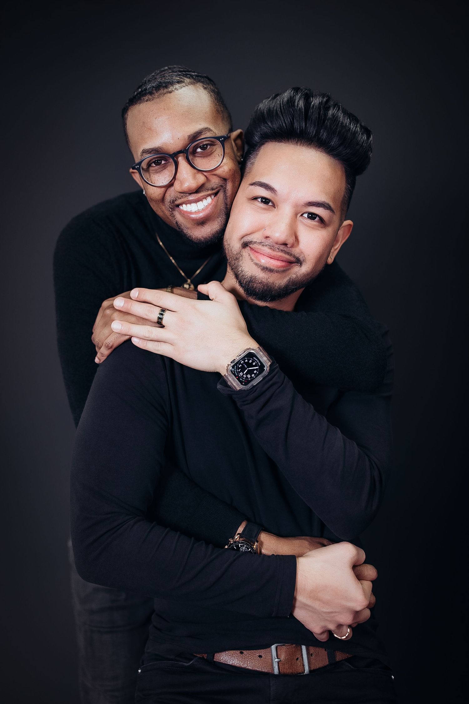 Two people in black outfits embrace and smile warmly against a dark studio backdrop in a series of intimate photos.