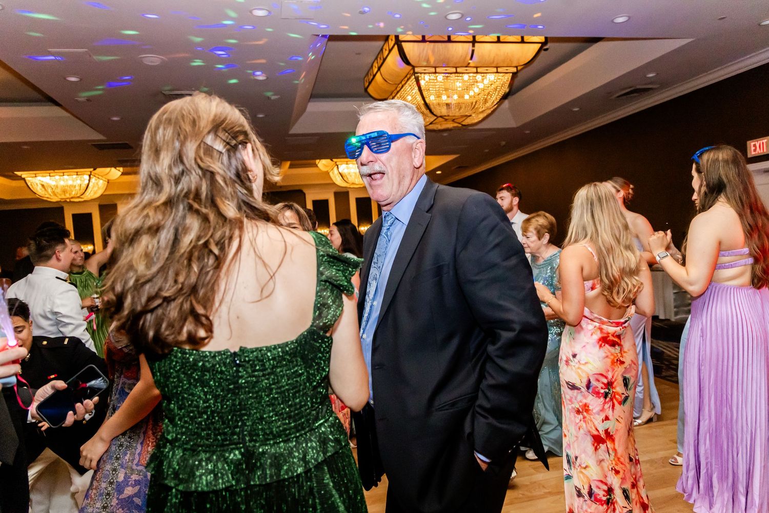 People dancing and socializing at an elegant ballroom event with crystal chandeliers and formal attire.