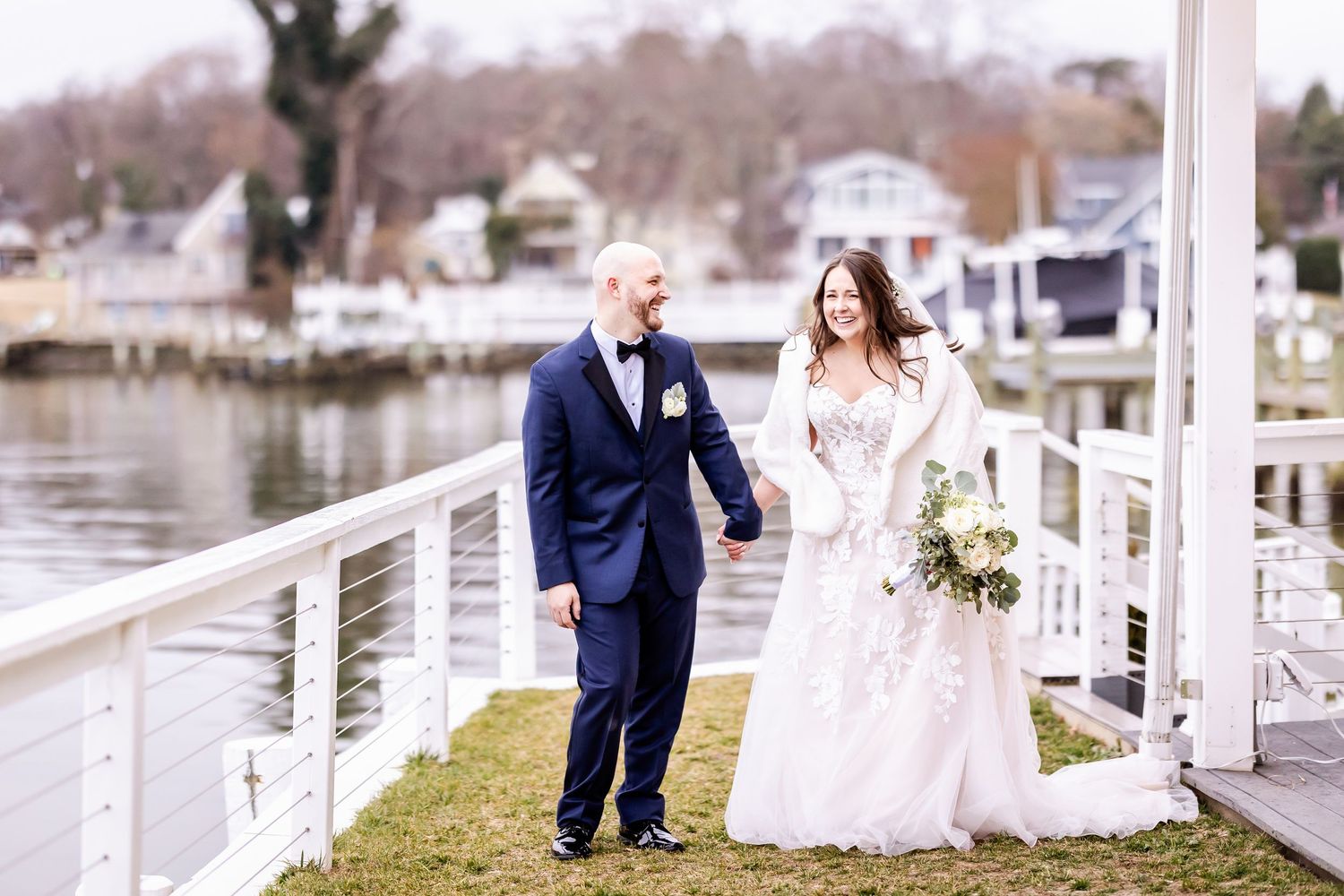 A couple in wedding attire walks along a lakeside dock on an overcast winter day.