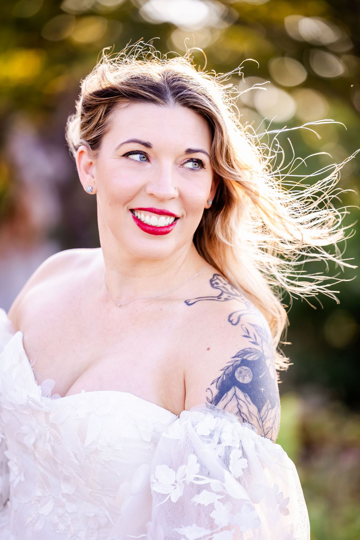 A bride in a white dress smiles joyfully outdoors with windswept hair and red lipstick in a natural setting.