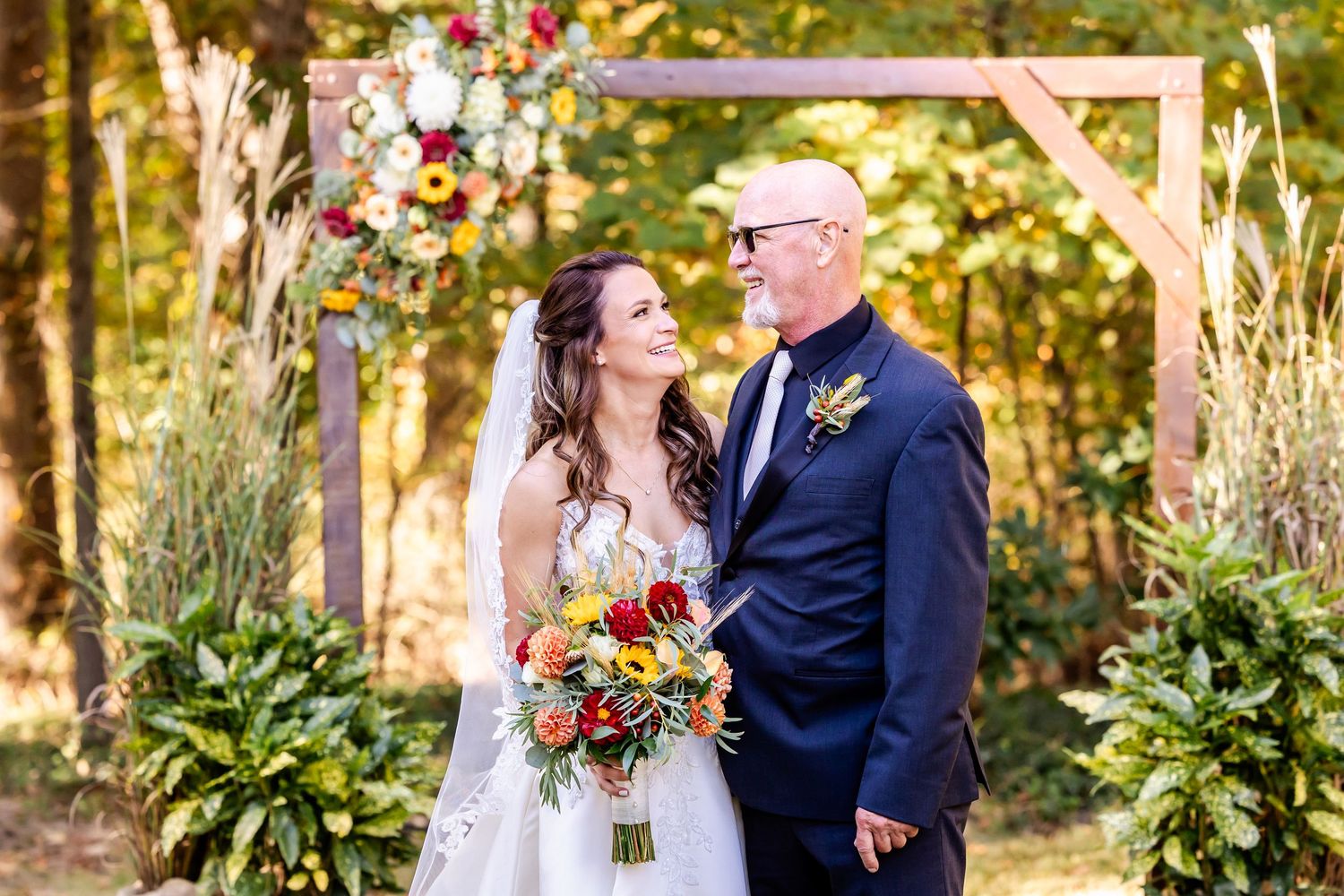 Autumn outdoor wedding ceremony with colorful floral arch and natural woodland backdrop.