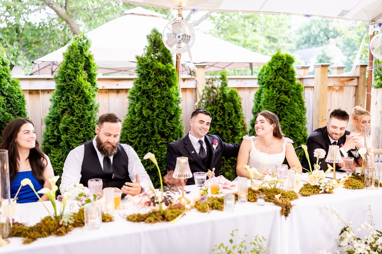 Wedding party seated at long tables decorated with greenery under a white tent.
