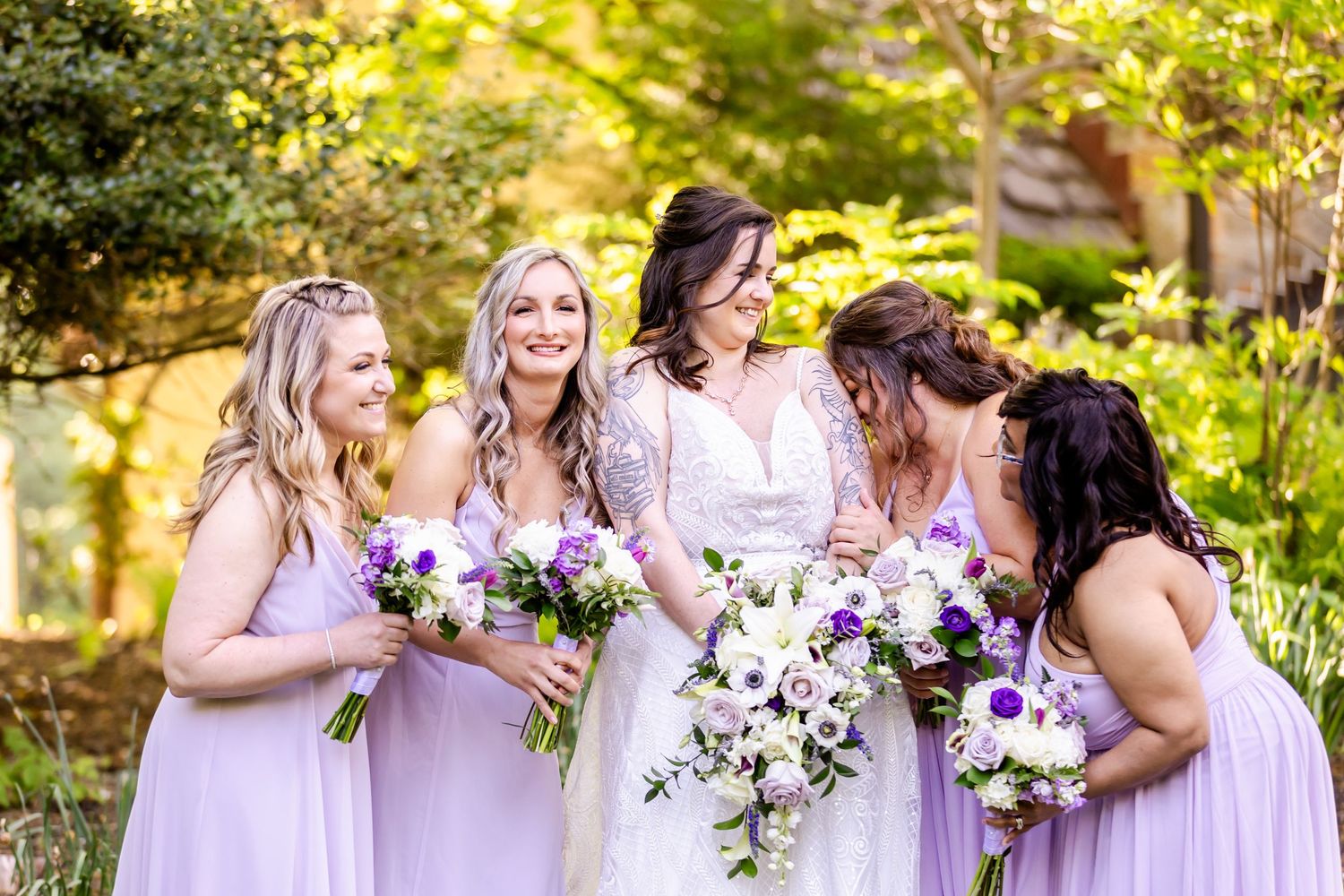 Wedding party in light purple dresses sharing a joyful moment while holding beautiful floral bouquets in a garden setting.