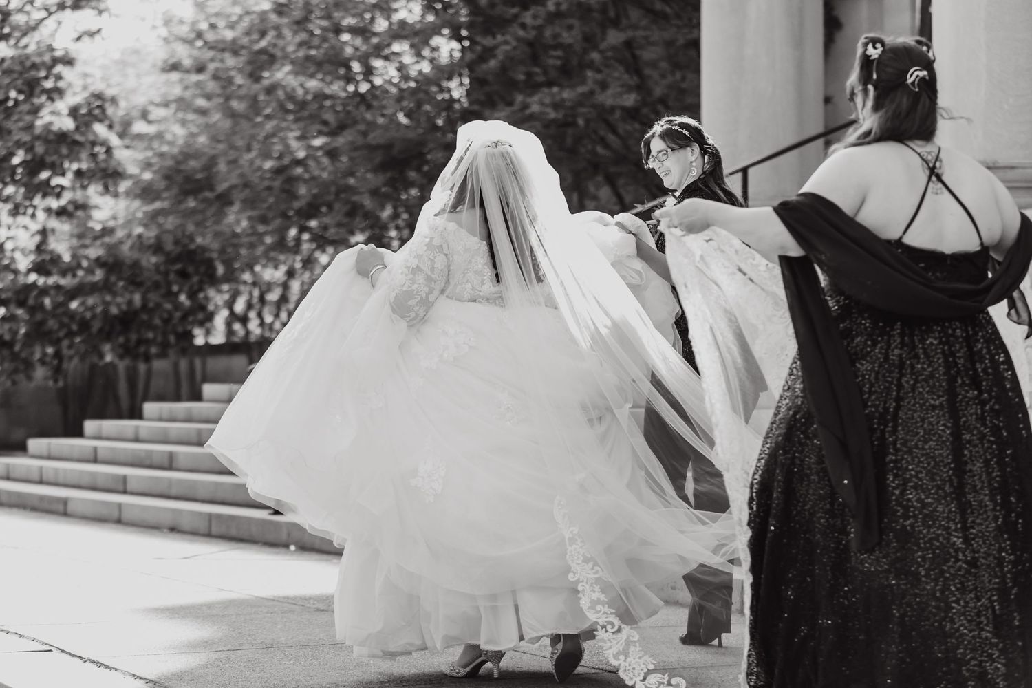 Black and white photo capturing attendants helping with bride's flowing veil on outdoor steps.