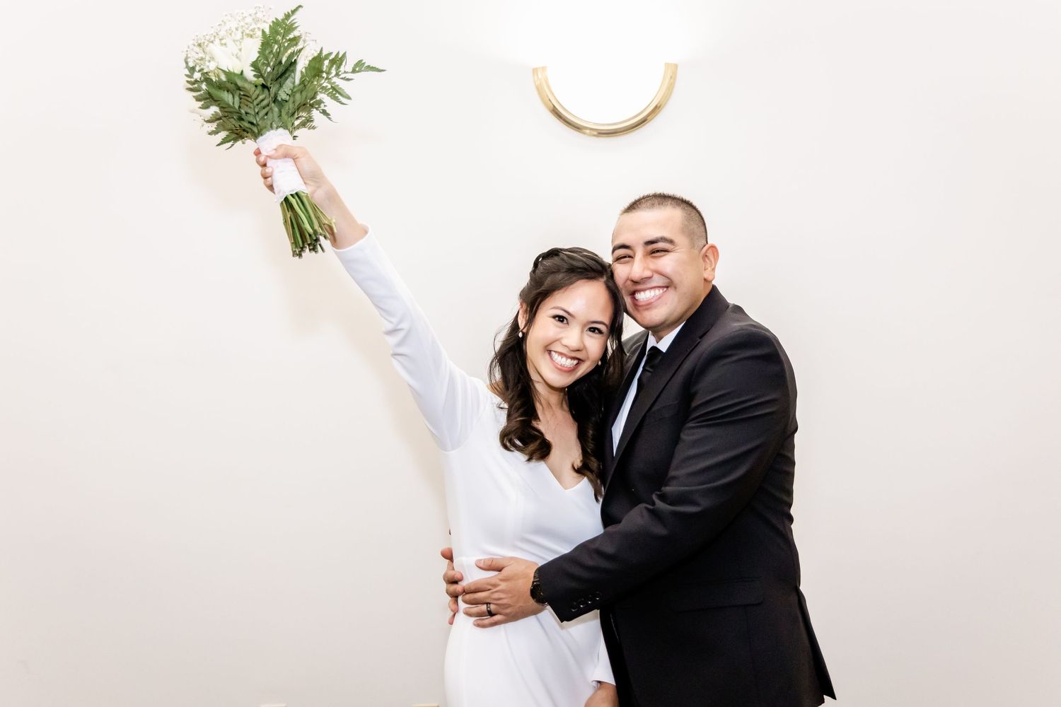 Newlyweds celebrate with bride holding bouquet high in victory pose against white wall.
