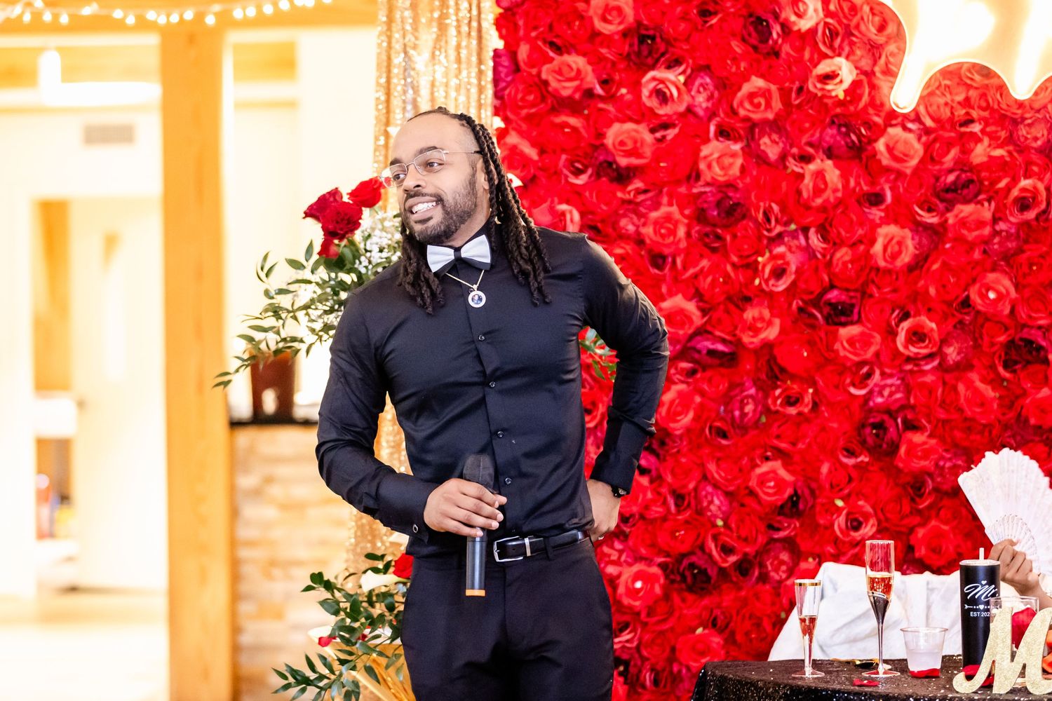 Speaker or performer in black attire presenting against dramatic red rose wall backdrop.