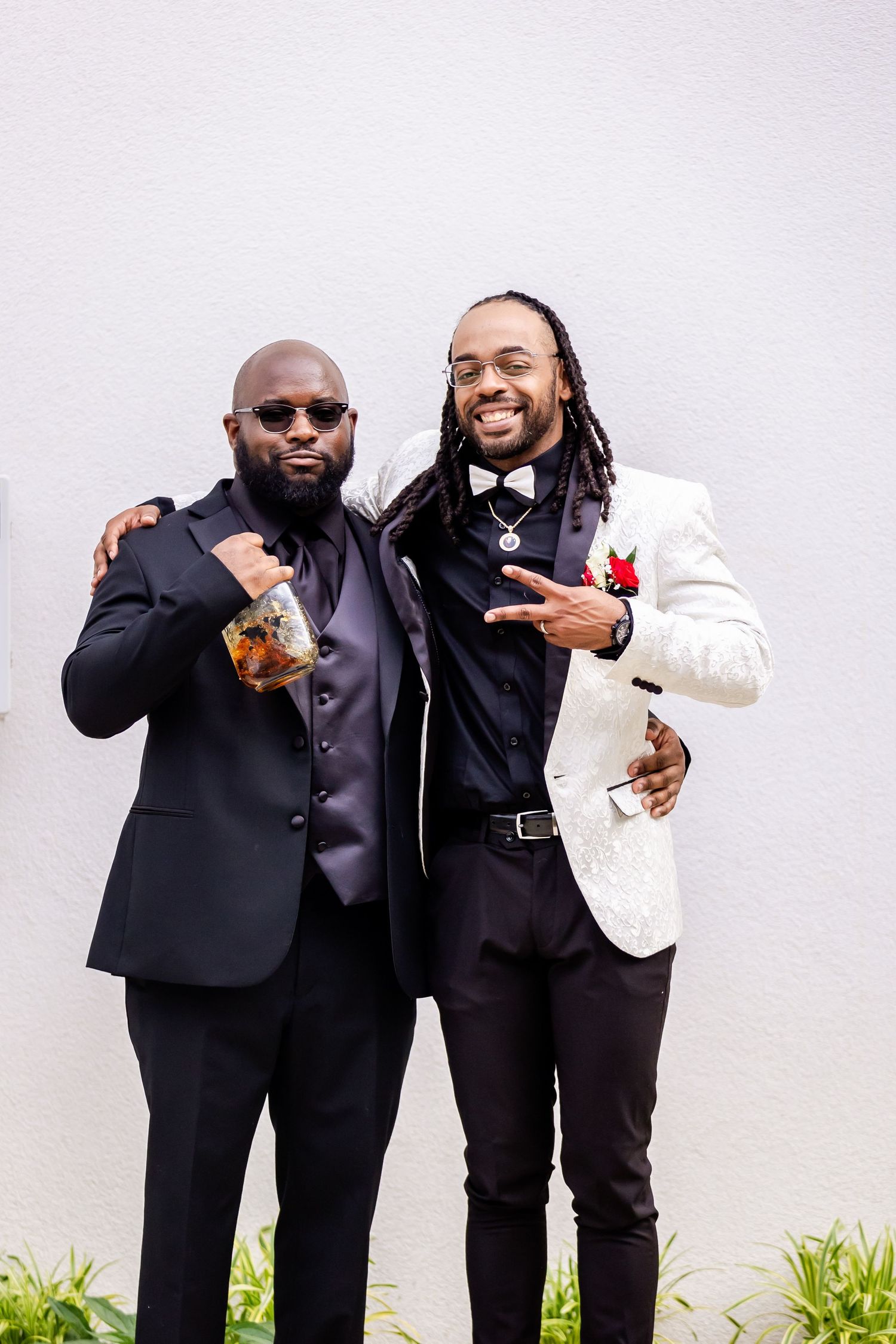 Two stylishly dressed men share drinks and pose against white wall.