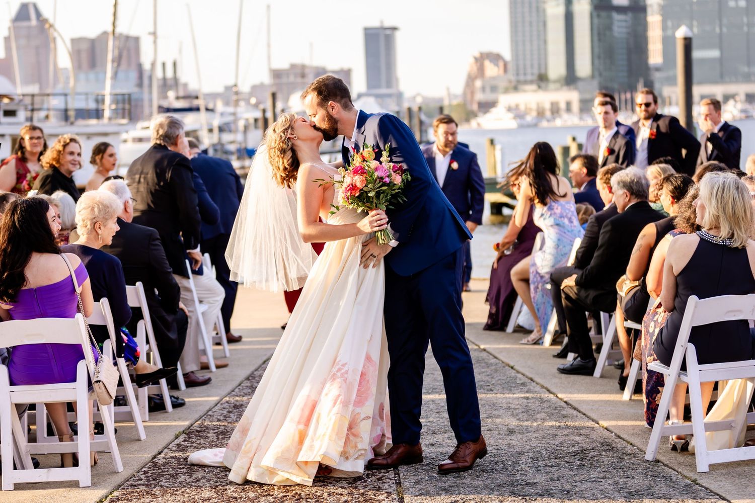 A wedding couple shares their first kiss during an outdoor ceremony with guests seated on white chairs by the marina.