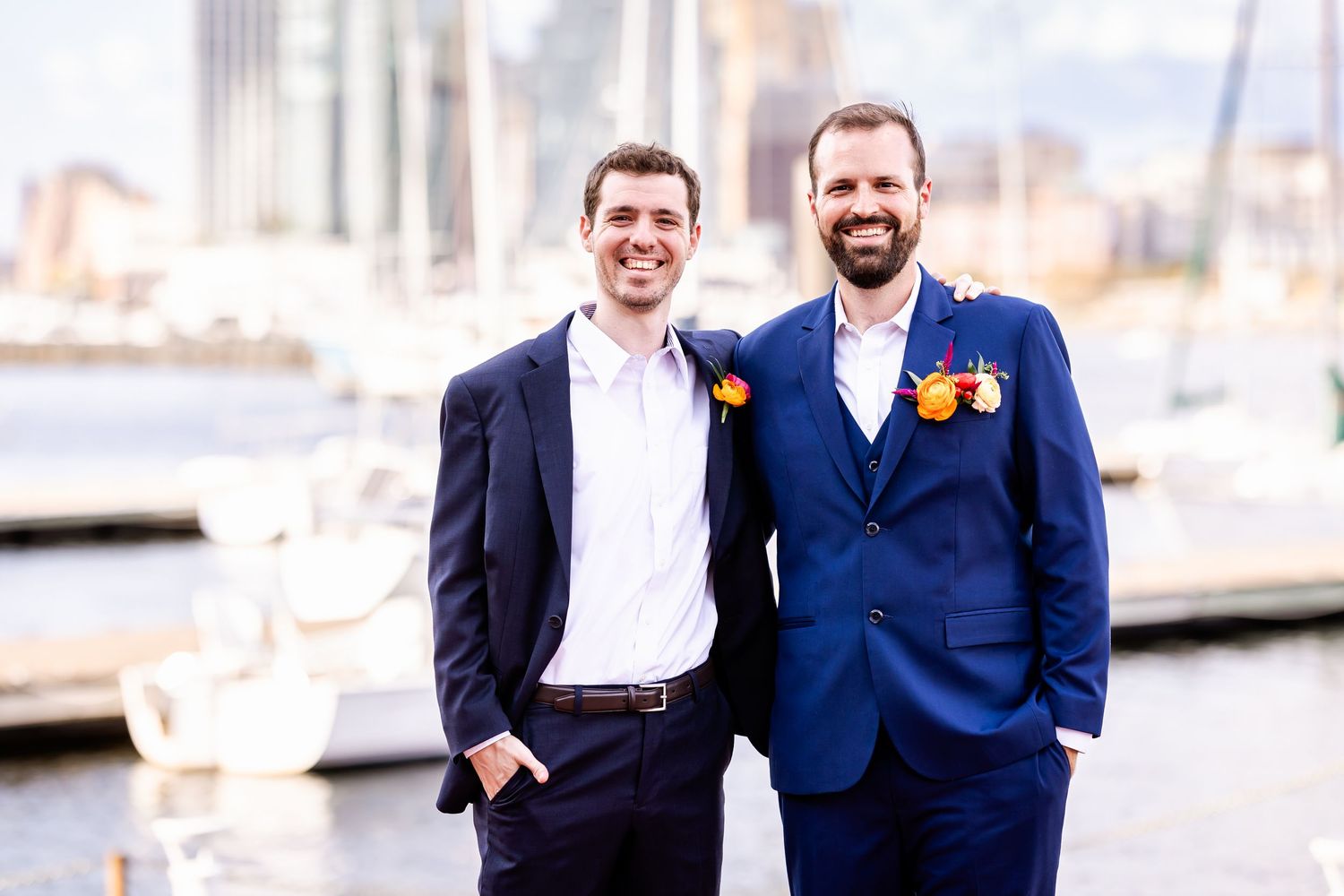 Two people in suits smile while standing on a dock with boats and buildings behind them.