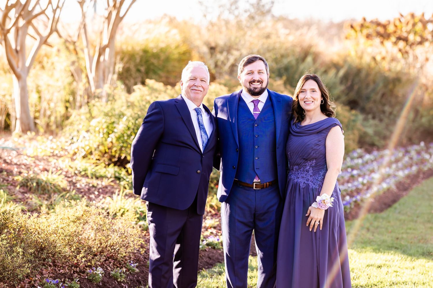 Family group in coordinating navy and purple formal attire pose together in an outdoor garden setting.