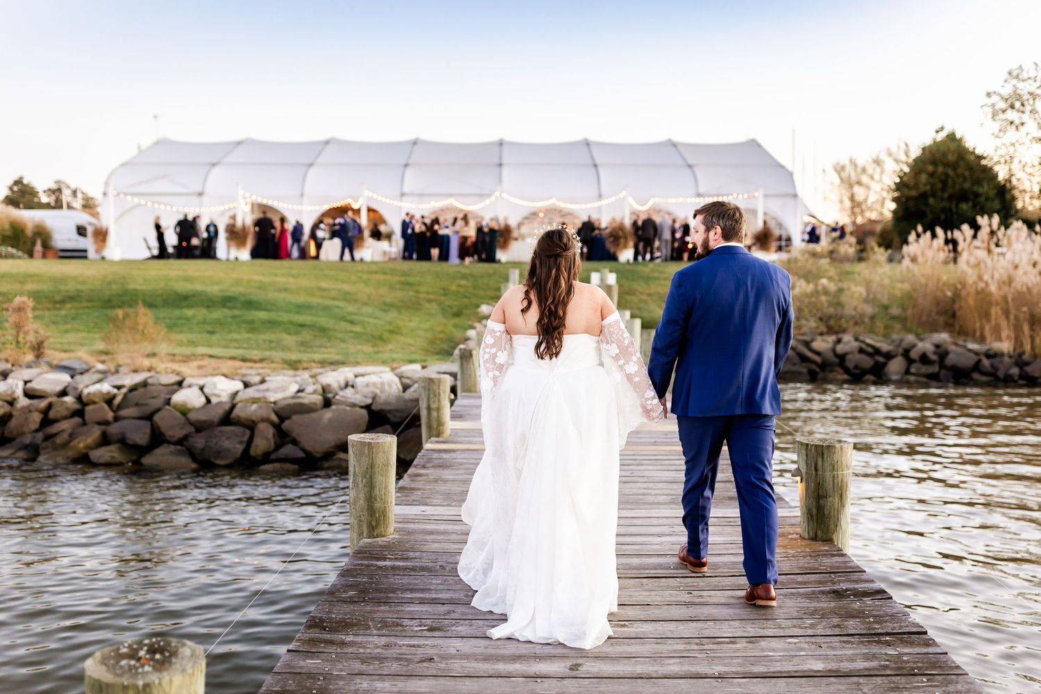 A wedding couple walks down a pier towards their tented reception by the water.