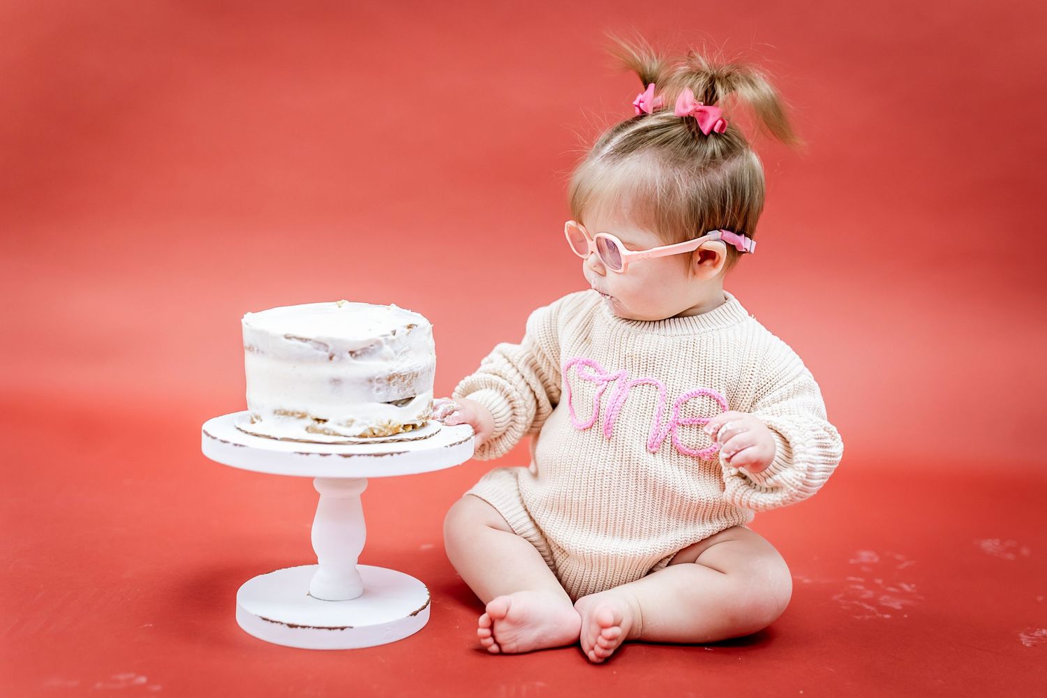 Baby reaches for white cake on pedestal against red backdrop.