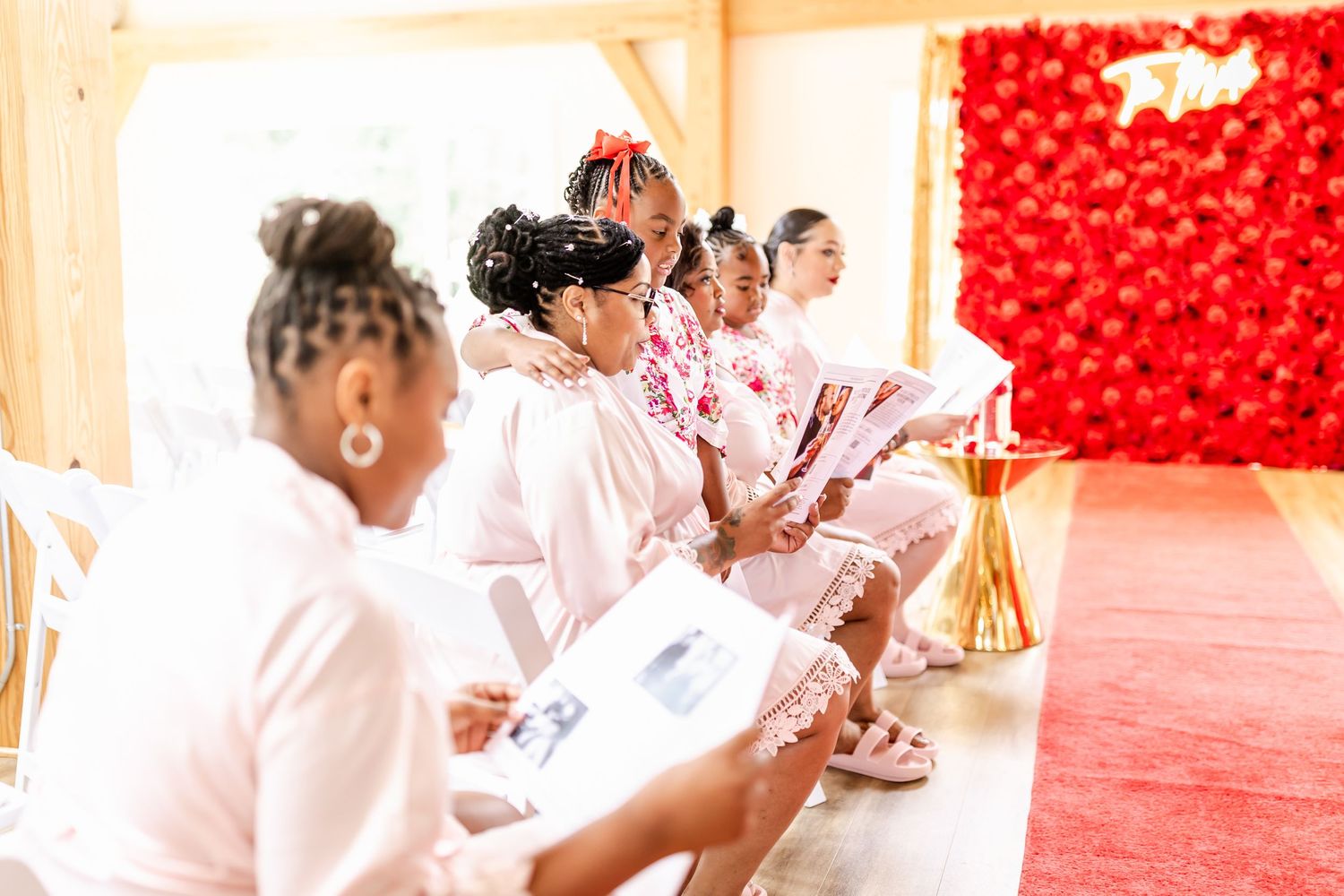 Wedding party members in pink robes seated in rows with a red backdrop and gold decor.