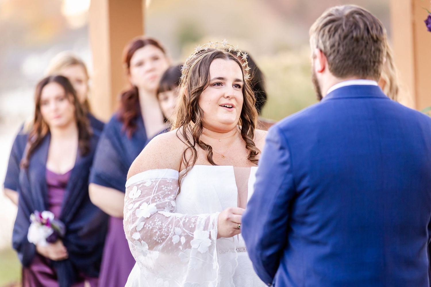 A bride in a white lace dress laughs joyfully during her wedding ceremony outdoors.