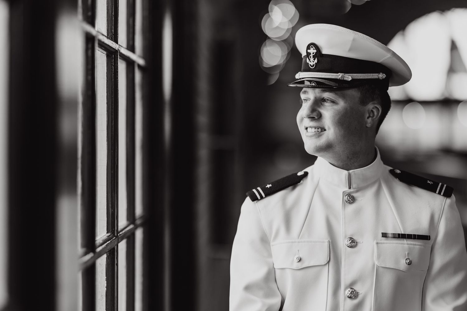 Naval officer in white dress uniform looking pensively through window in black and white portrait.