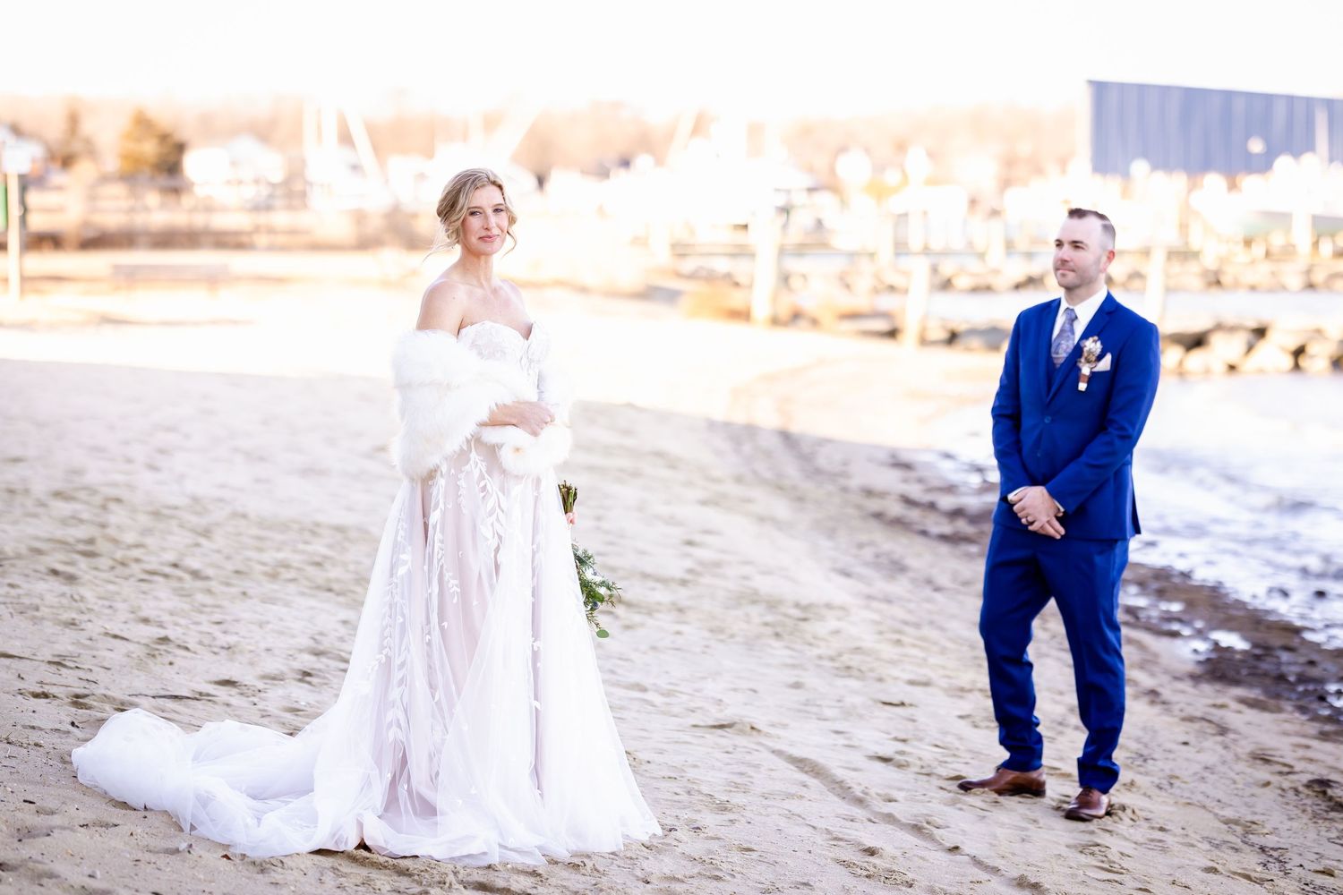 A bride in a lace gown and groom in navy suit pose separately on a beach during sunset.