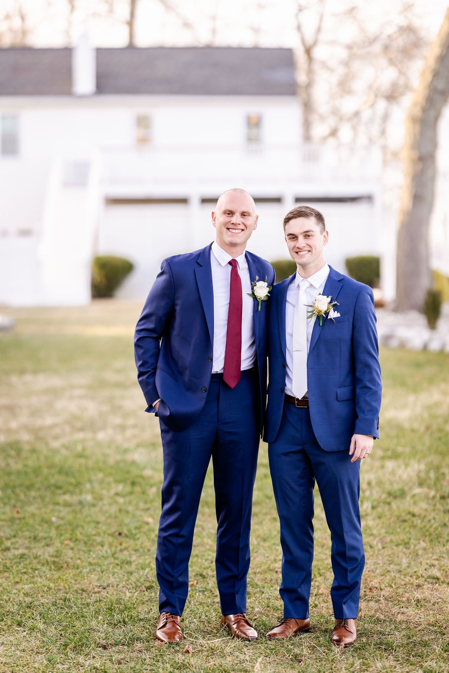 Two people in navy blue suits and brown shoes stand together on grass in matching wedding attire.
