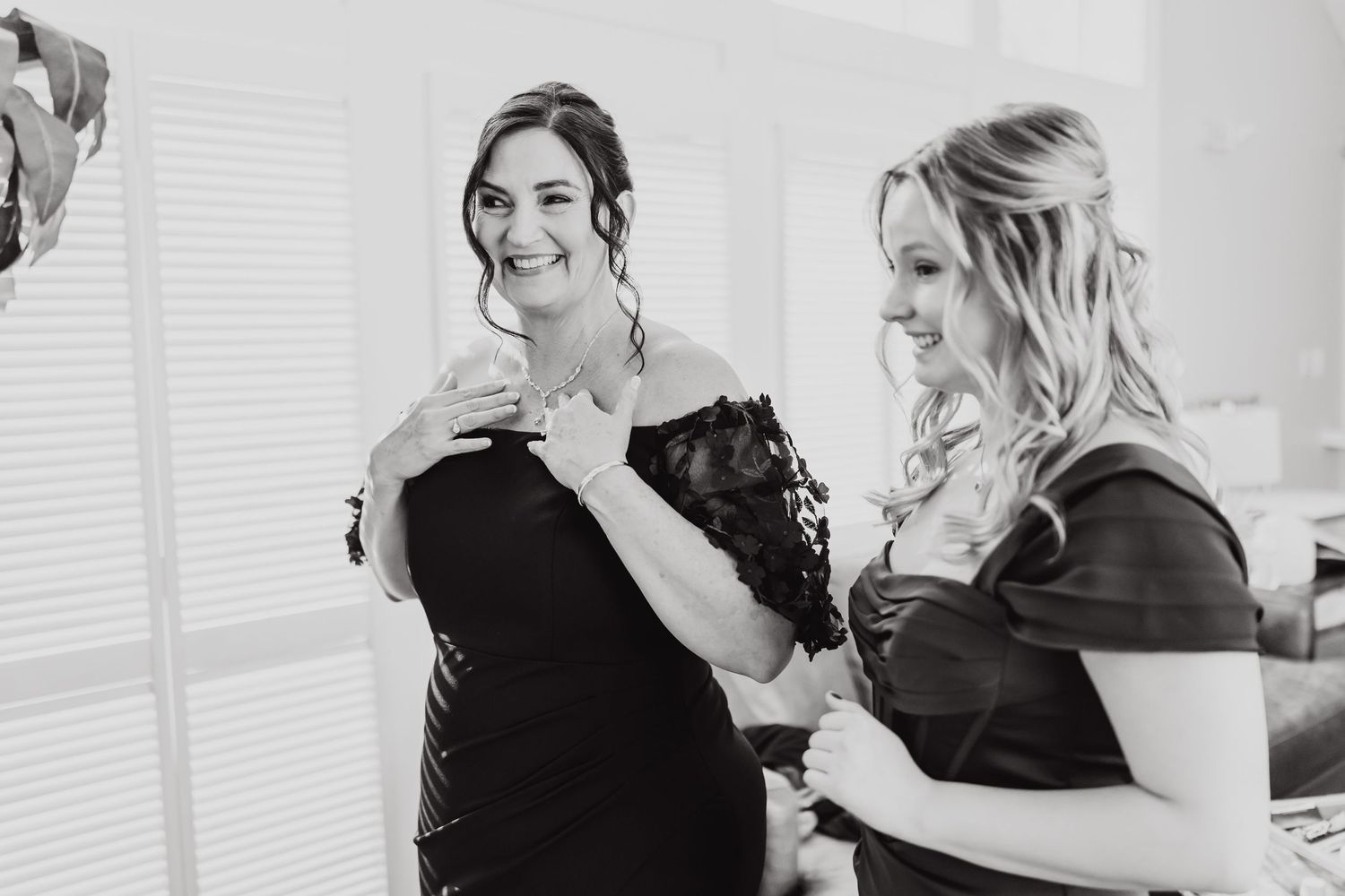 Two women in dark dresses share joyful moment during wedding preparations.