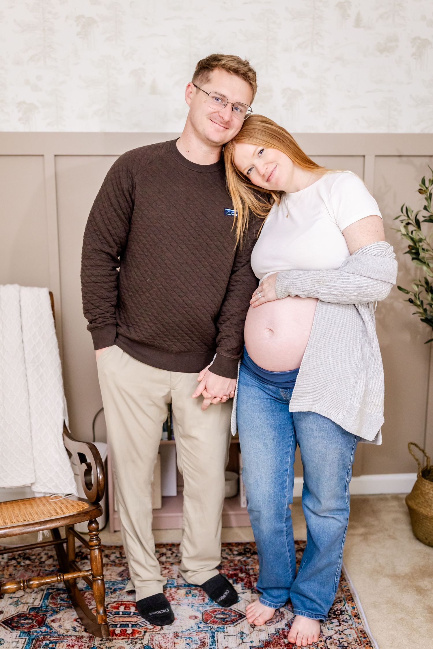 An expectant couple in casual attire shares a tender moment while standing in their cozy living room.