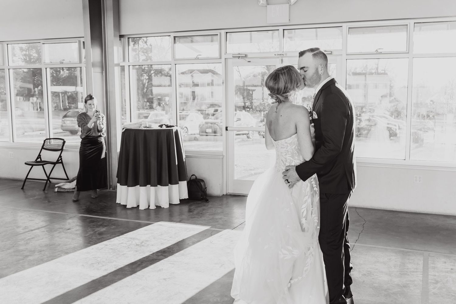 A couple shares their first dance at a wedding reception in a bright sunlit room with large windows.