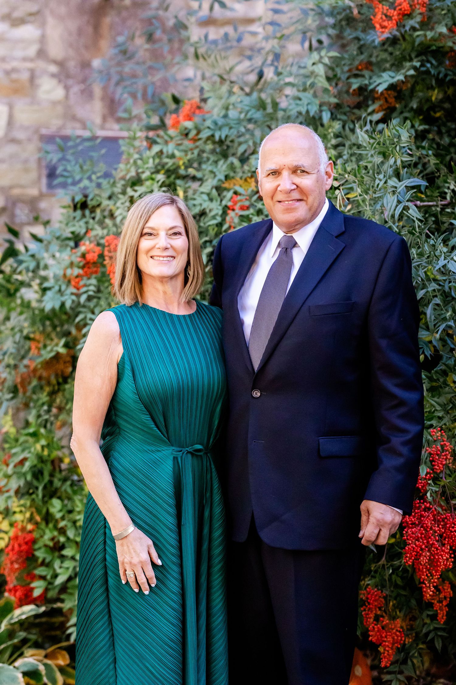 Elegant couple poses against stone wall with vibrant red flowers, wearing formal evening attire.
