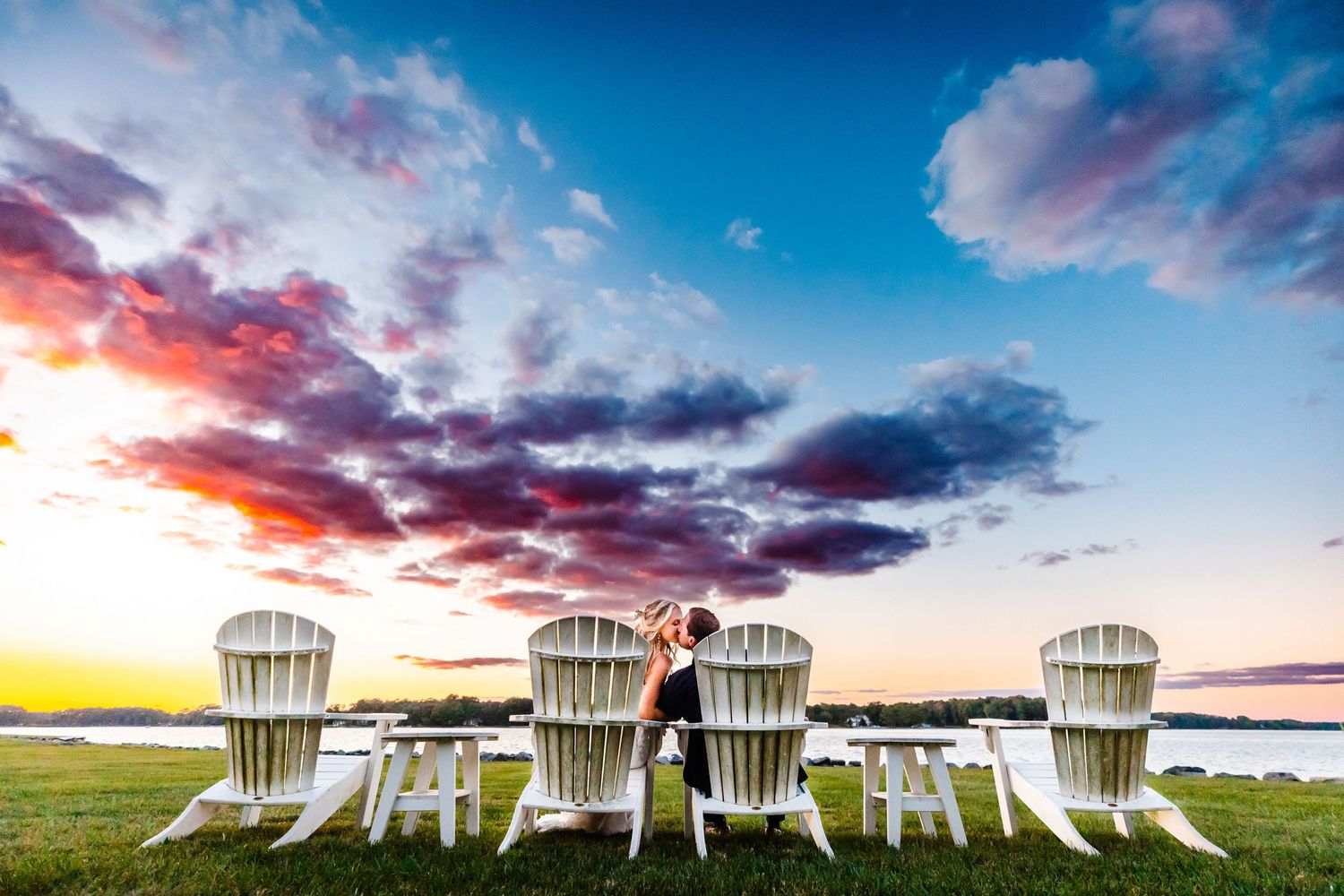 Adirondack chairs line a grassy waterfront at sunset with dramatic pink and purple clouds in the sky.