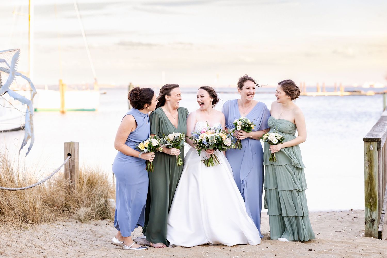 A bridal party wearing green and blue dresses poses together on a sandy beach with bouquets.
