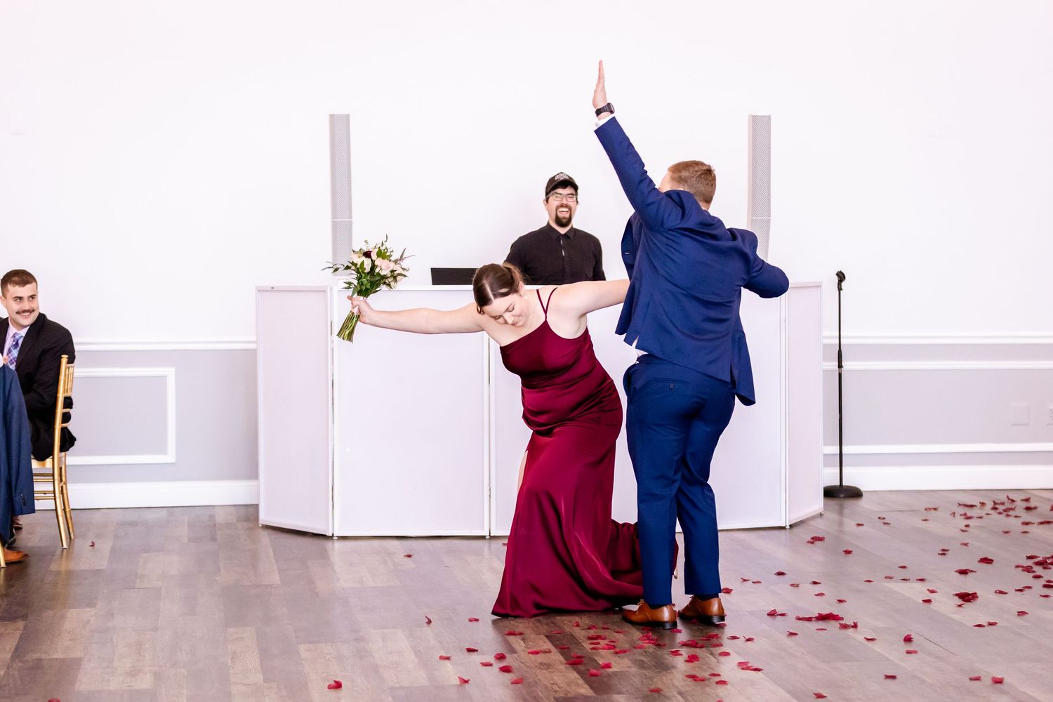 Wedding party celebrates with energetic dance moves in white reception venue.