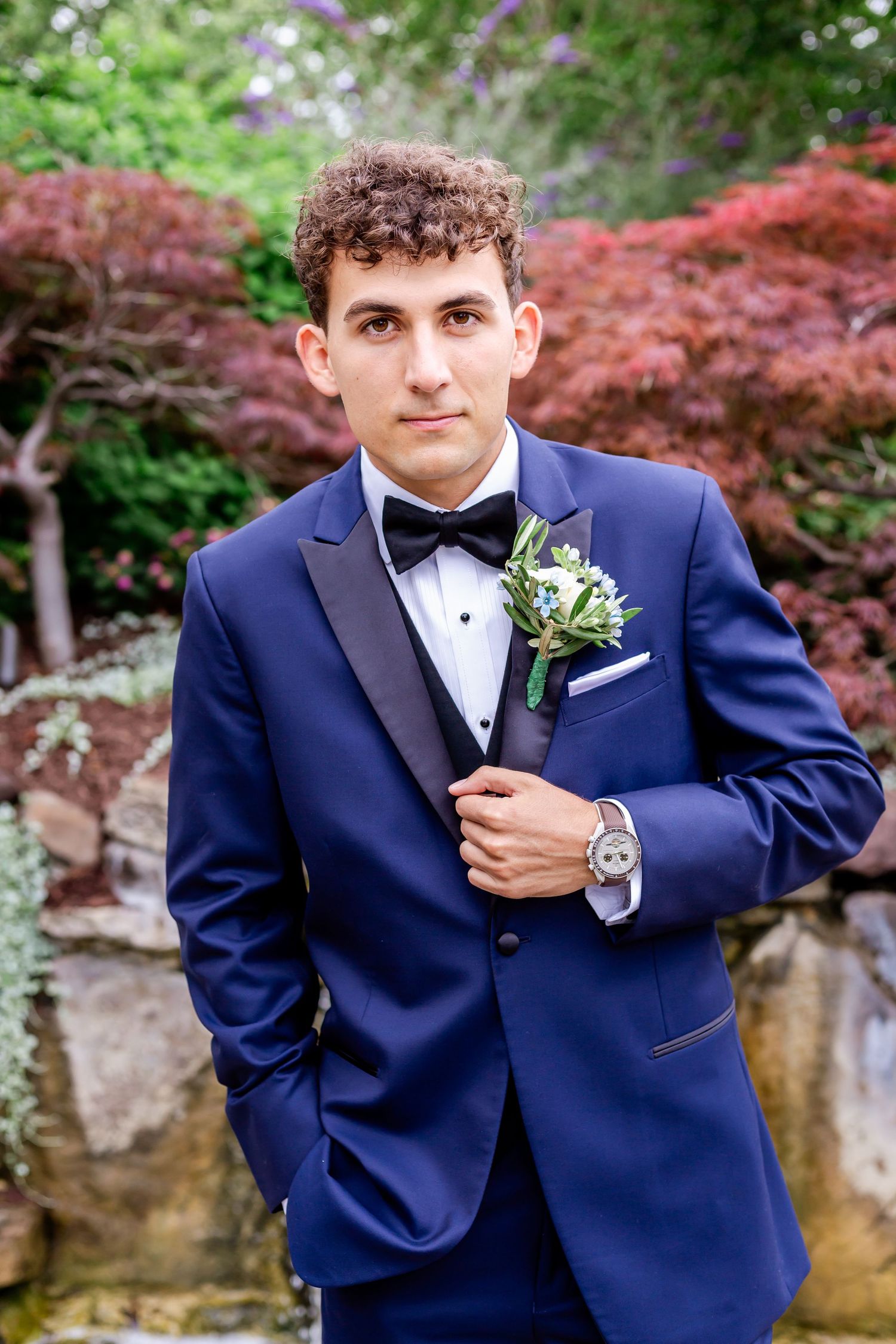 A groom in a navy blue tuxedo poses against a backdrop of vibrant red Japanese maple trees.
