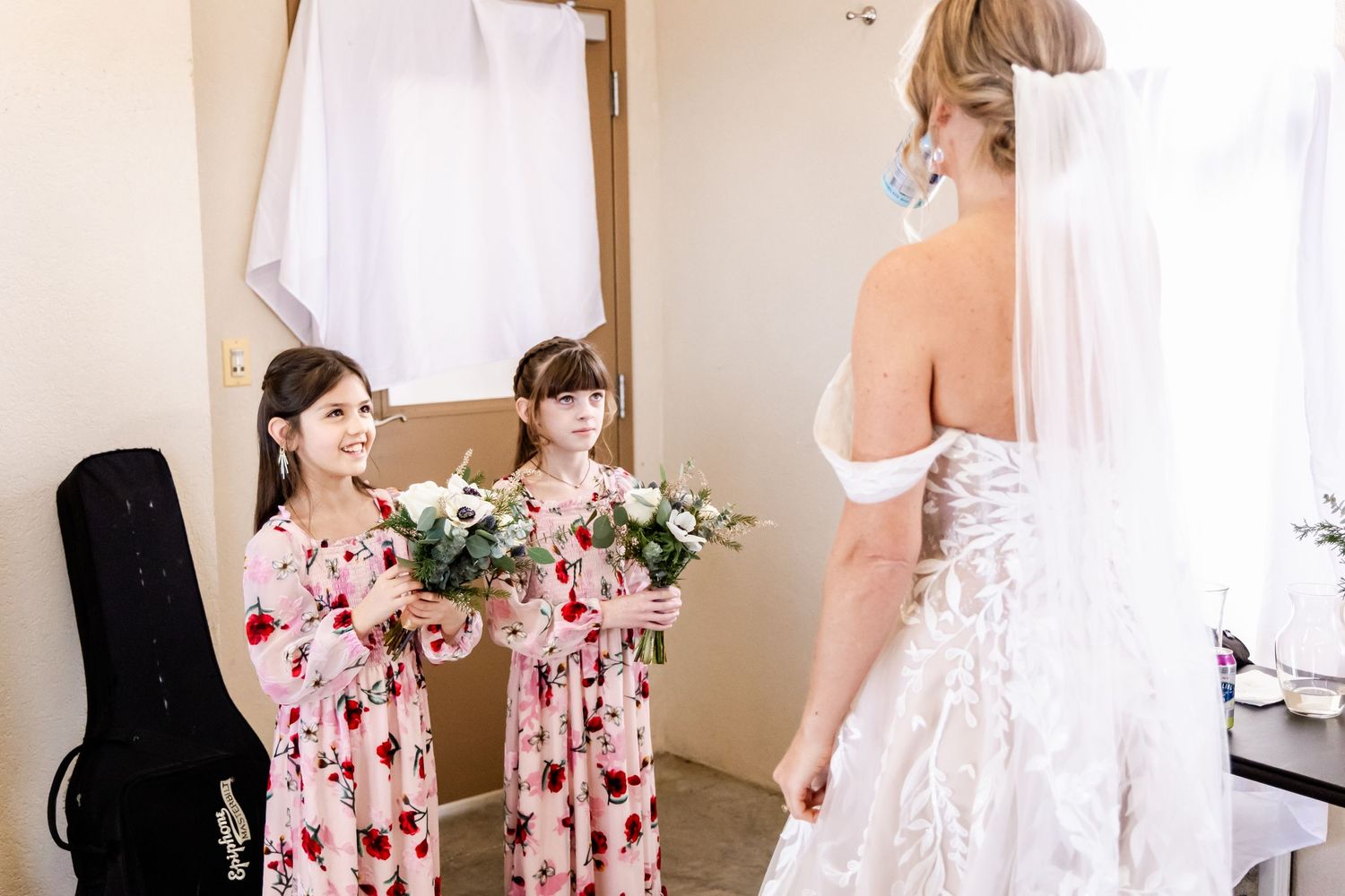 Young attendants in pink floral dresses admire a bride in her wedding gown.