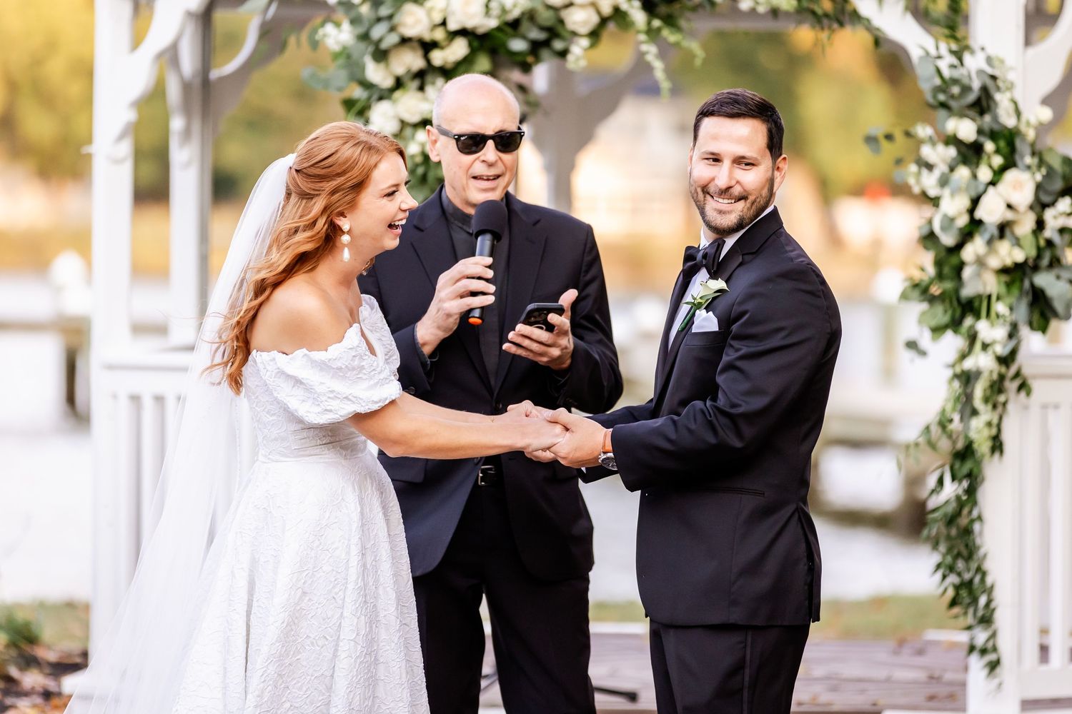 A beautiful outdoor wedding ceremony takes place under a floral arch with sunlit greenery in the background.