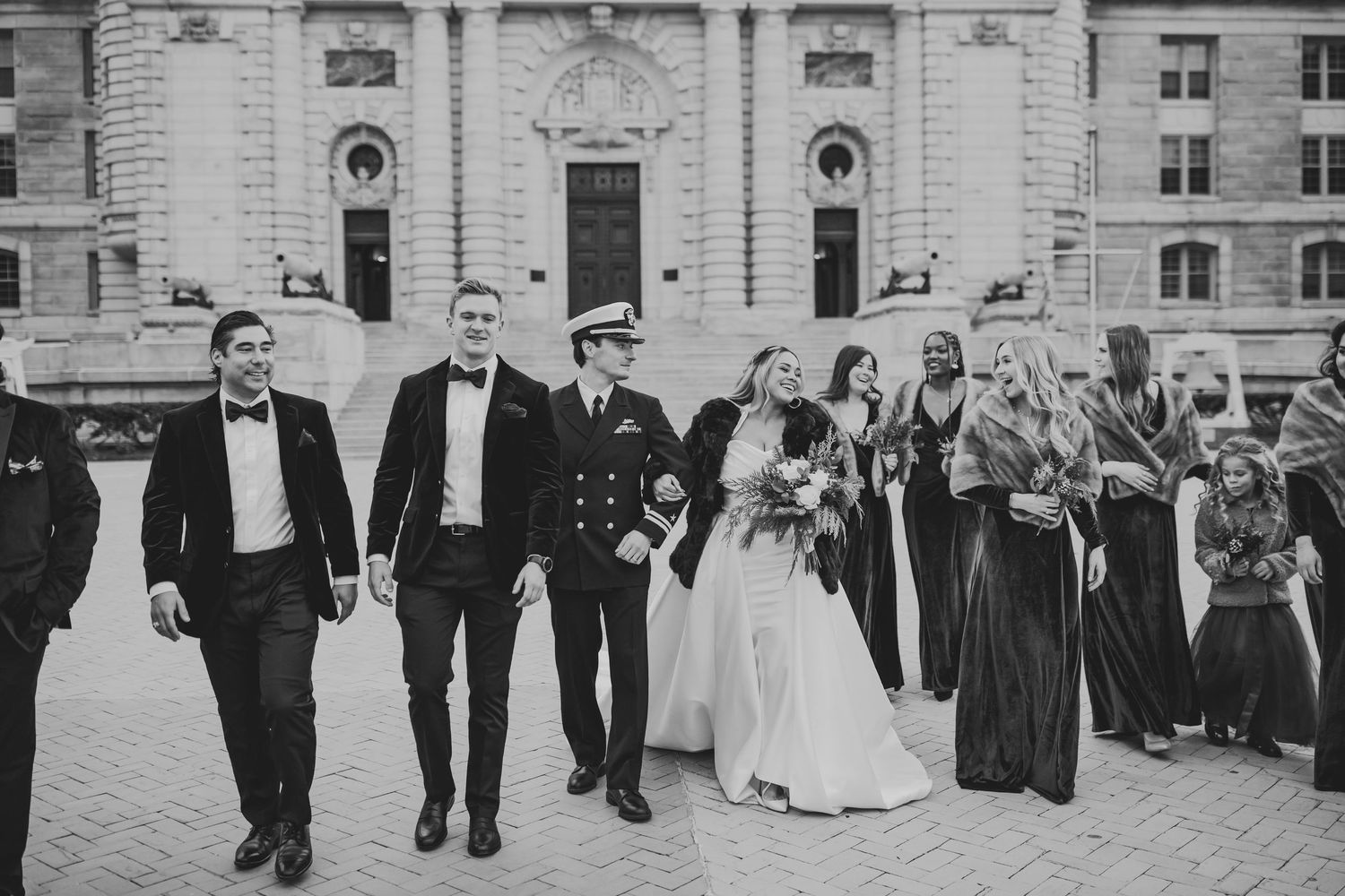 Wedding party walks together outside historic building in black and white photo with bride in white gown and formal attire.