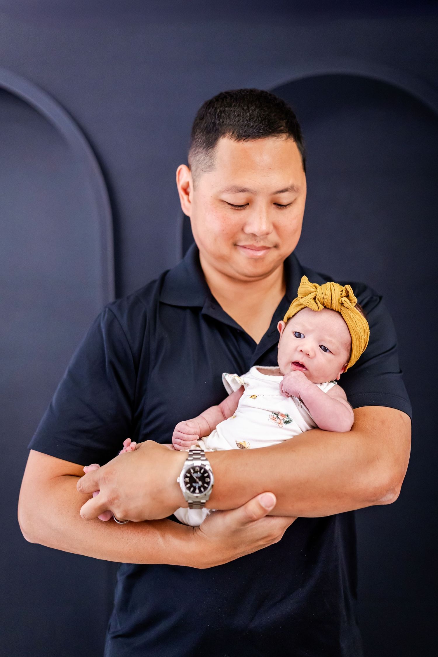Sweet portrait series showing an infant wearing a yellow bow headband being cradled in arms against a dark backdrop.