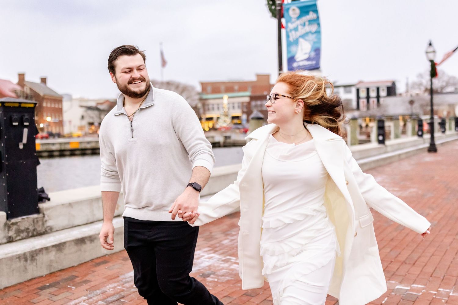 A couple joyfully walks together on a brick sidewalk in a city during winter, wearing light colored sweaters.
